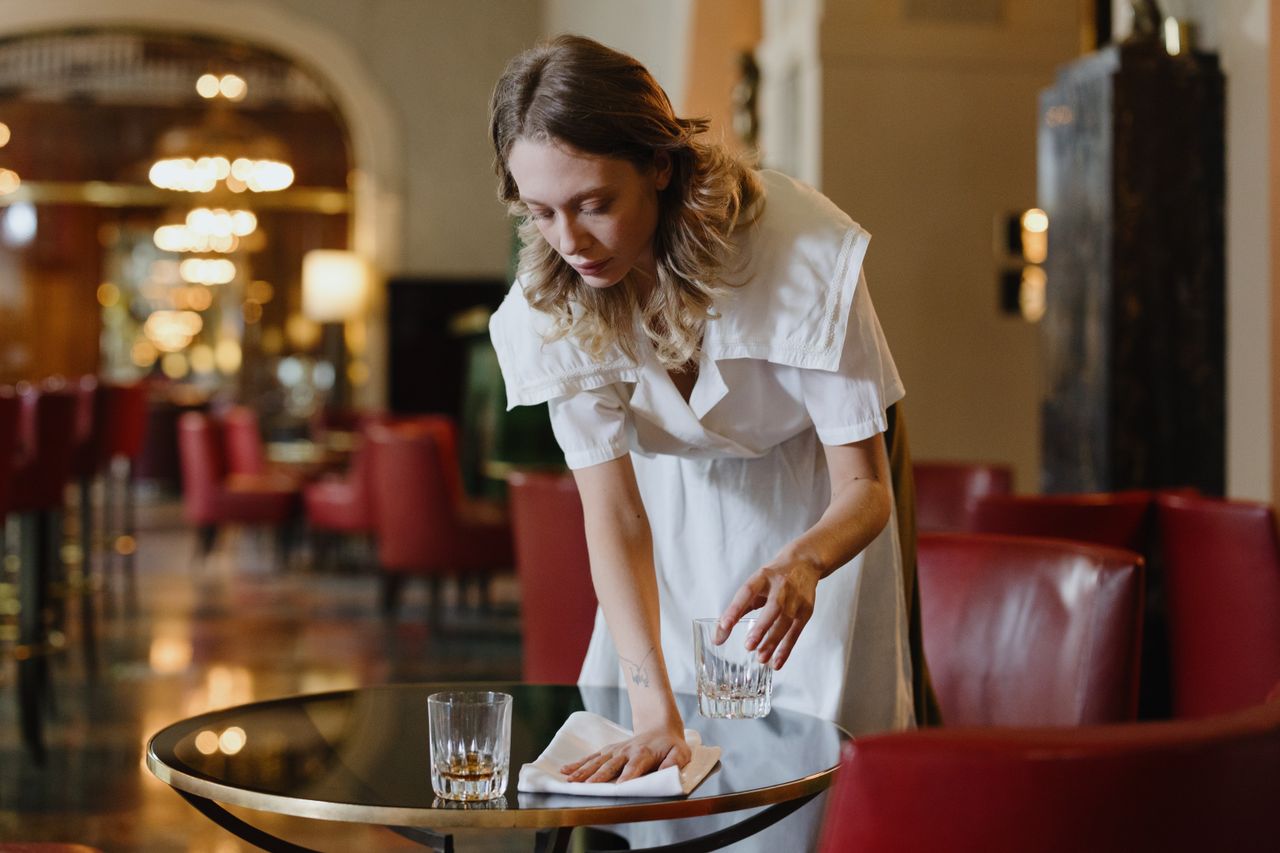 A waitress in a white dress wiping a table in a stylish restaurant. Warm, inviting atmosphere.